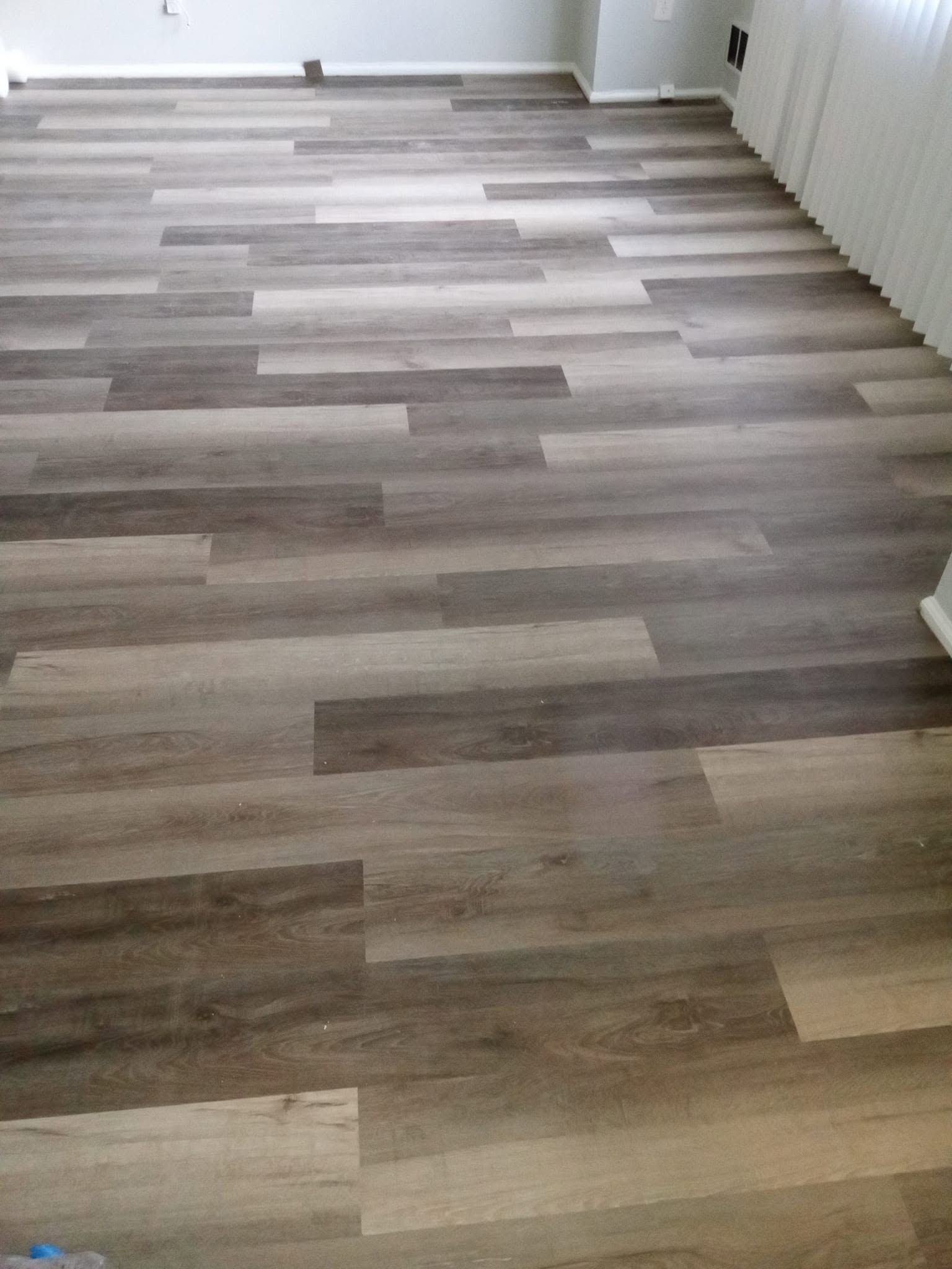 Empty room with gray wood-look flooring and a white wall, lit by natural light.