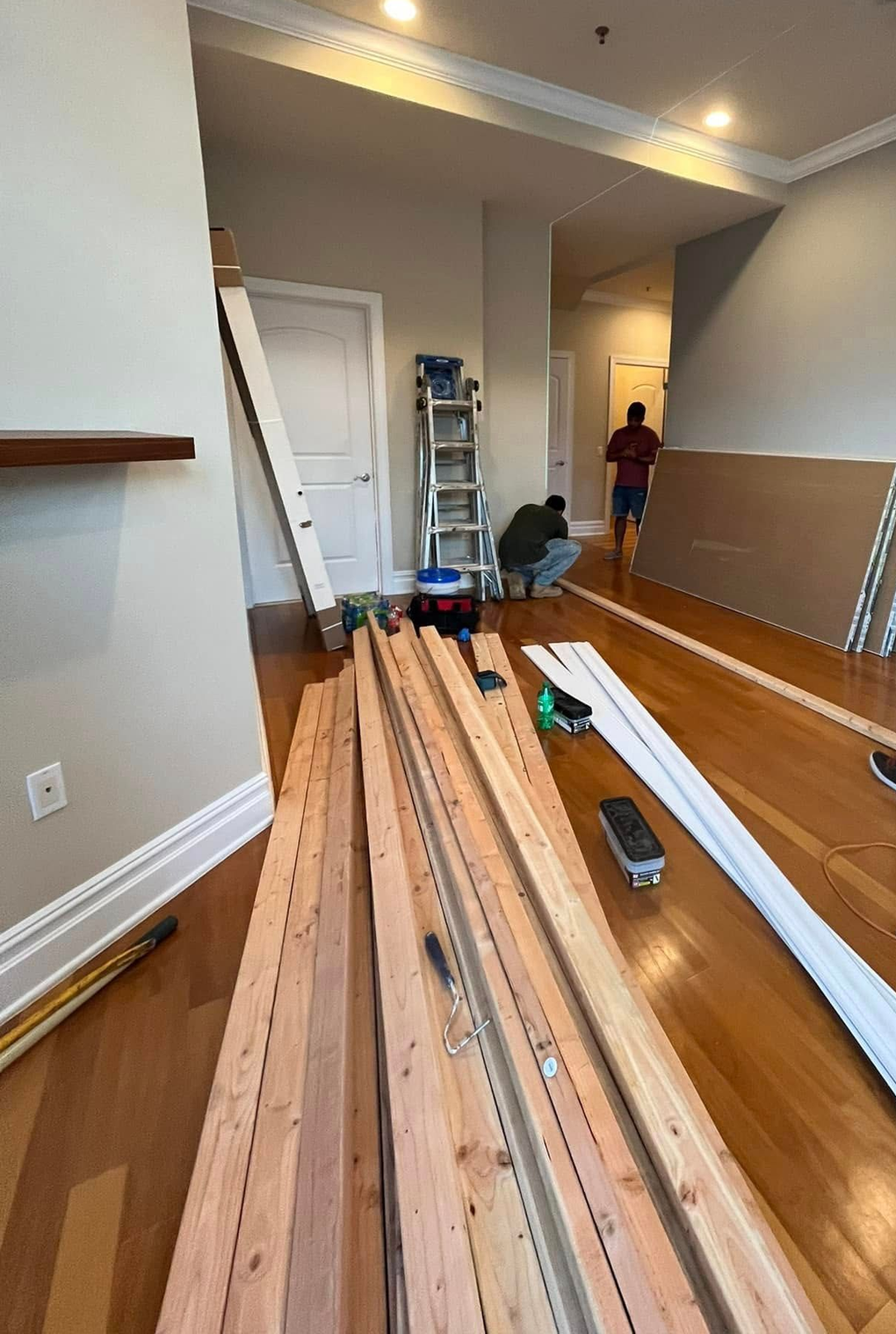 Wood planks and tools on a hardwood floor during home renovation, with a ladder and a person in the hallway.