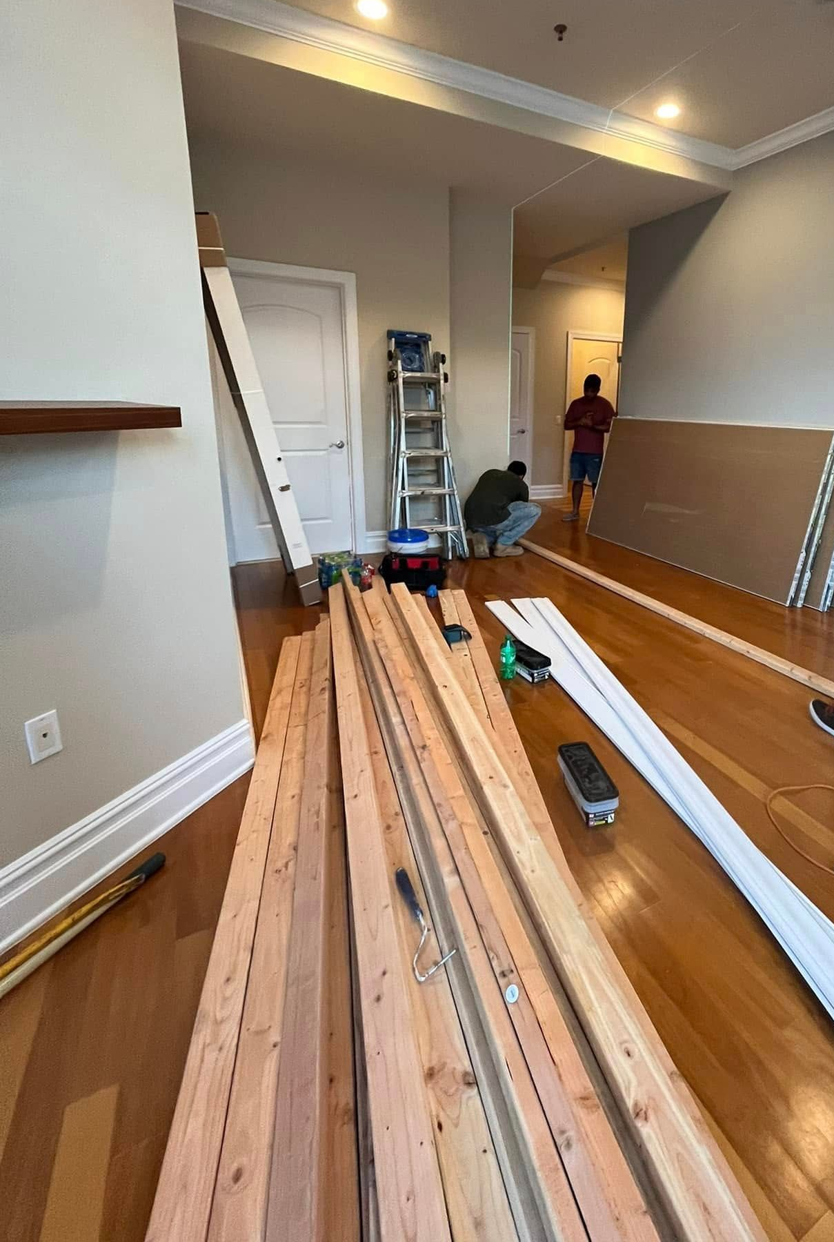 Interior room with hardwood floors, stacked lumber, tools, and a person in the doorway during renovation.