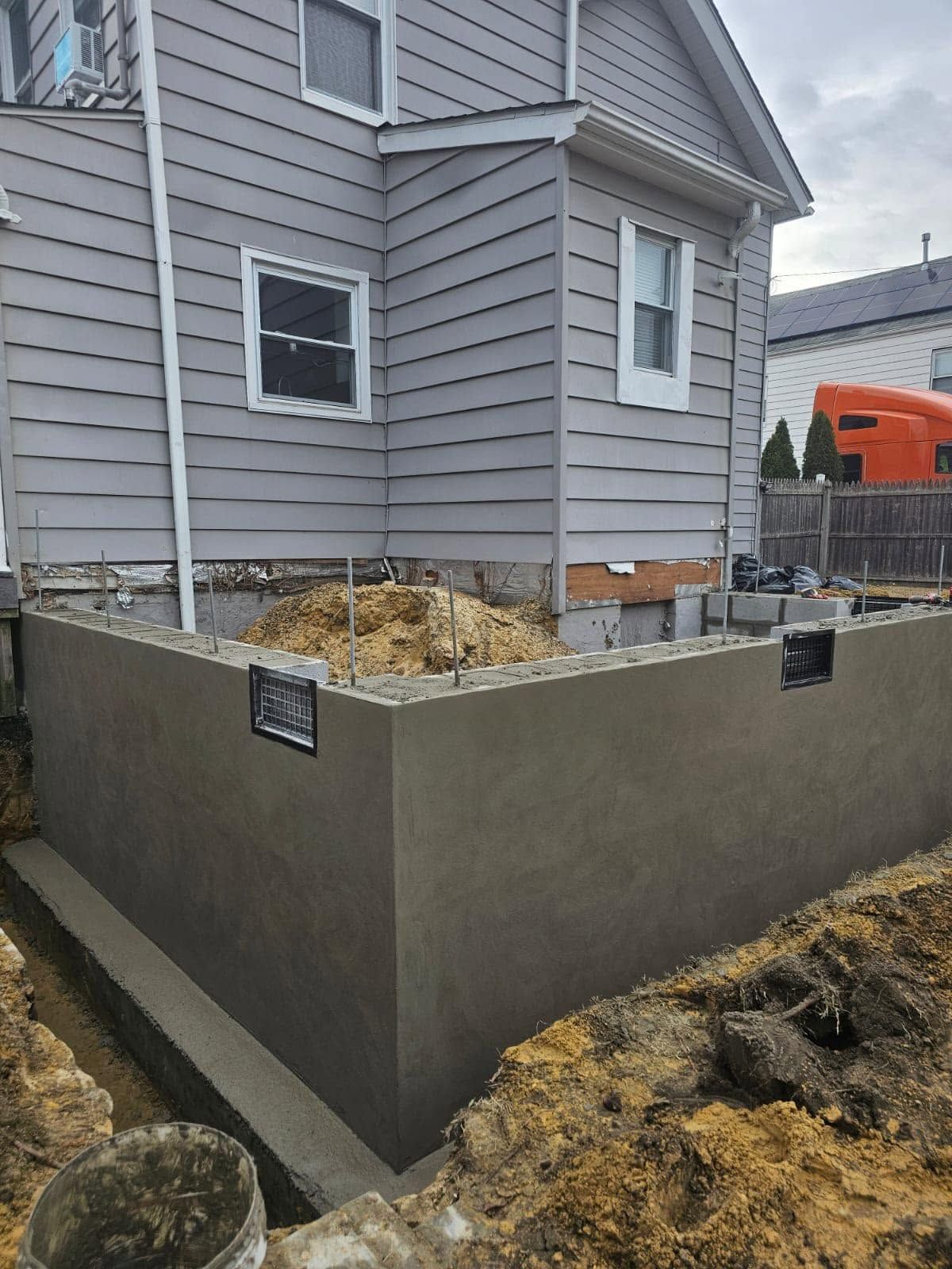 Gray house with a new concrete retaining wall and an orange excavator in the background.