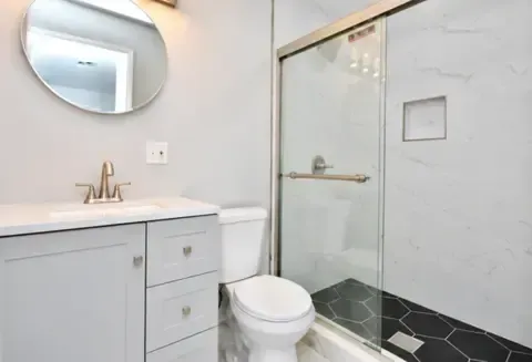 Bright bathroom with vanity, round mirror, toilet, and glass shower with black tile floor.