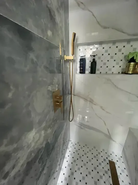 Modern shower with gray marble tiles, gold fixtures, white basin, and small mosaic floor accents