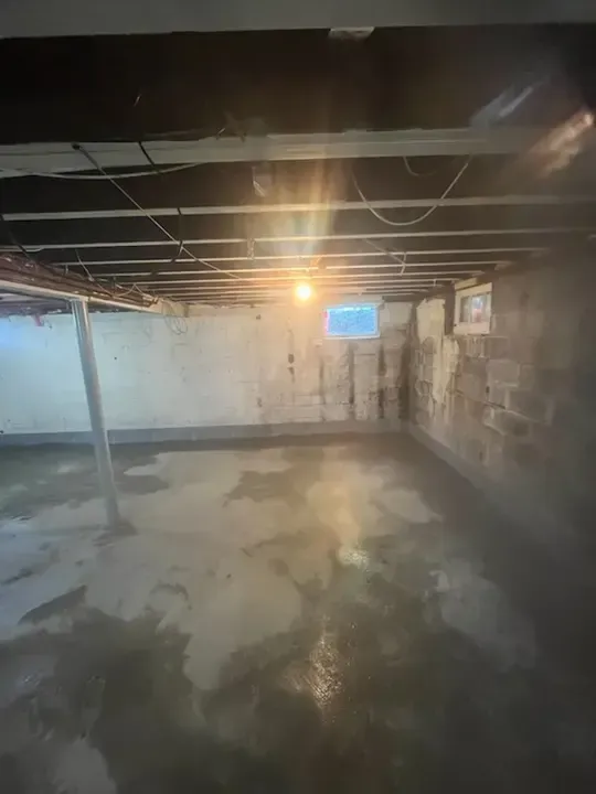 Empty basement with exposed ceiling beams, concrete floor, and a small blue window on the back wall.