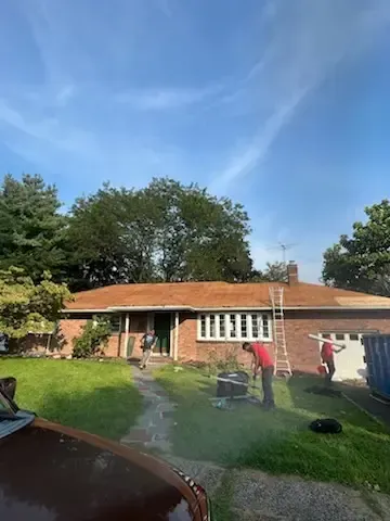 People in a sunny backyard, one watering a lawn near a single-story house with a red roof.