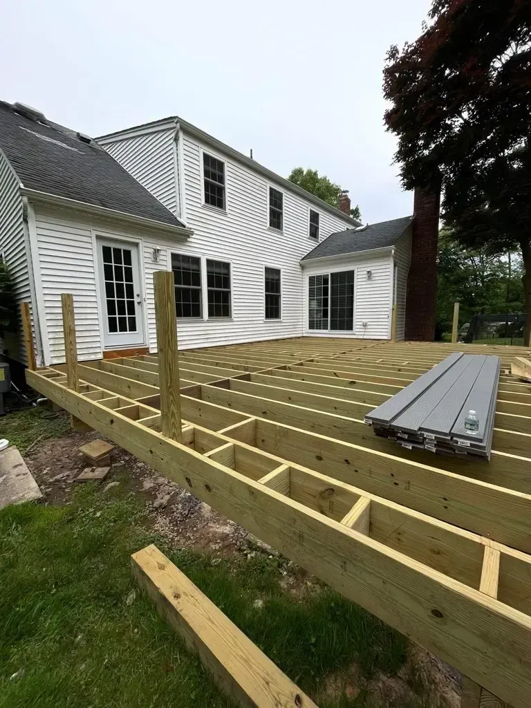 Backyard deck framing under construction beside a white house, with lumber and joists visible.