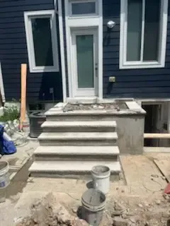 Concrete front steps and stoop under construction outside a blue house, with buckets and tools in the foreground