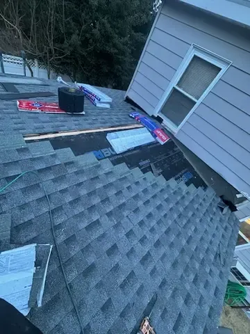 Damaged roof with missing shingles and scattered tarps, viewed beside a house wall and window