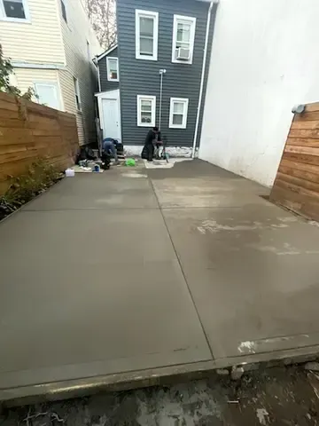 Concrete driveway beside a narrow alley, with a dark gray house and trash bins at the end.