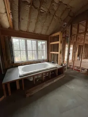 Unfinished bathroom with exposed framing, large window, and a bathtub set on a platform.