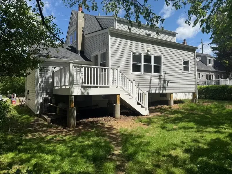 White house with a raised deck and stairs in a sunny backyard with trees and grass