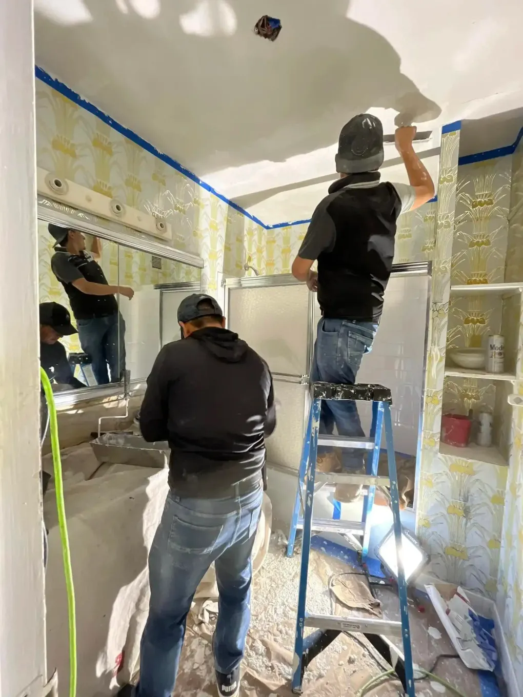 Two workers painting a bathroom, one on a ladder and one on the floor, with blue tape along the ceiling.