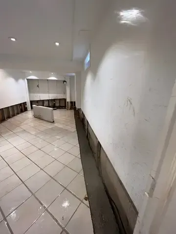 Empty white-tiled hallway with white walls and a low dark trim, viewed from a corner.