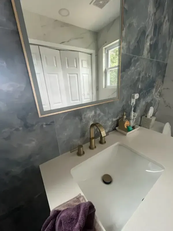 Modern bathroom vanity with white sink, brass faucet, dark marble walls, and a mirror reflecting a door.