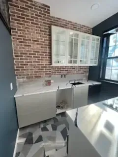 Modern kitchen with exposed brick wall, white cabinets, and gray lower cabinets beside a large window.