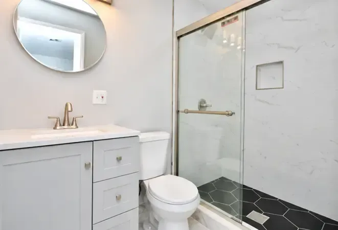 Modern bathroom with white vanity, round mirror, toilet, and glass shower with black tile floor