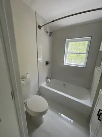 White bathroom with a toilet, bathtub, glass shower screen, and small window.
