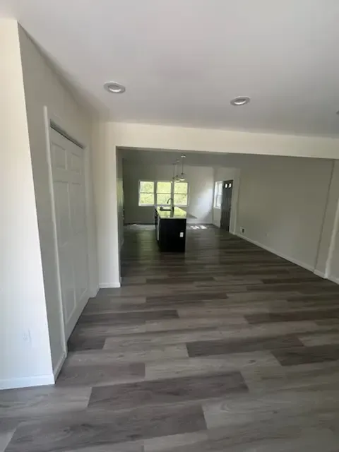 Empty modern living room with gray wood floors, white walls, recessed lights, and kitchen island in the distance