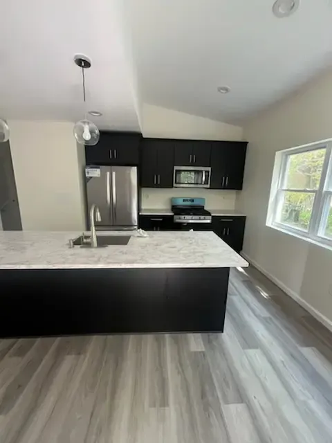 Modern kitchen with black cabinets, stainless steel appliances, marble island, and gray wood flooring.