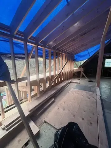 Roof framing under construction with exposed wooden rafters and blue tarps overhead