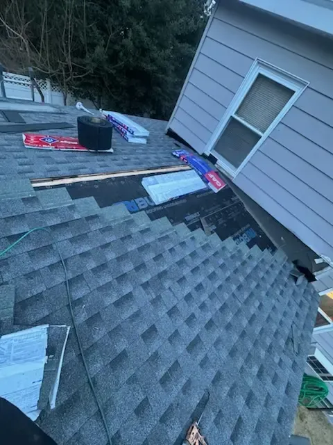 Roof with scattered shingles and tools beside a gray house wall, viewed from above