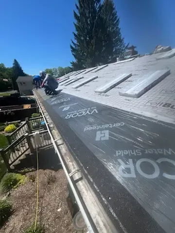 Two roofers installing black roofing felt on a sloped rooftop under a blue sky