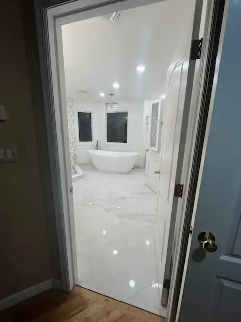 Bright white bathroom with freestanding tub, large mirror, and glossy tile floor, viewed through an open door