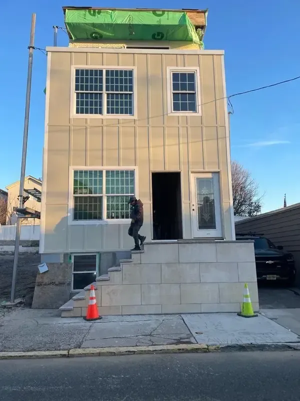 Person on front steps of a tan house under construction, with orange and green cones on the sidewalk.