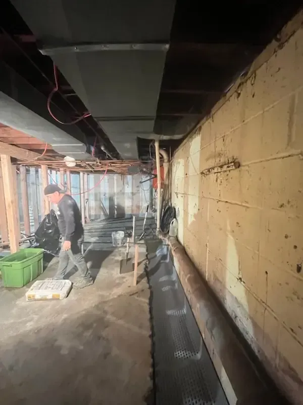Basement renovation site with worker, exposed wiring, and construction materials along a concrete wall.