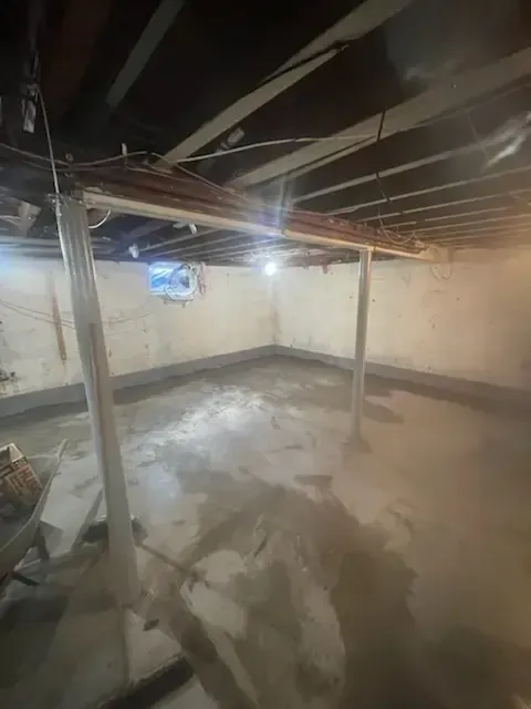 Dim unfinished basement with bare beams, concrete floor, support posts, and a small window.