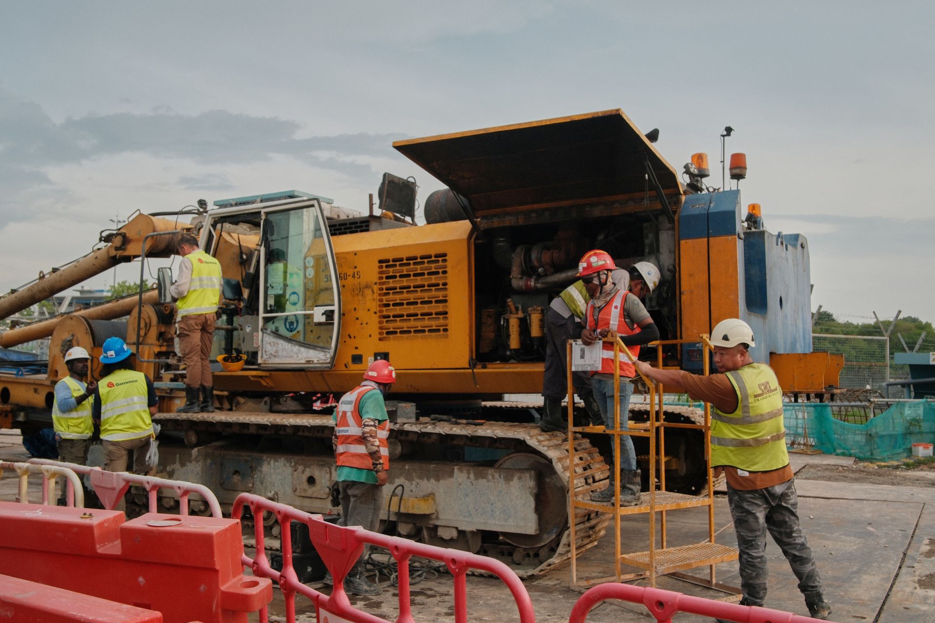 Construction crew working on a large orange drilling machine behind red barriers outdoors