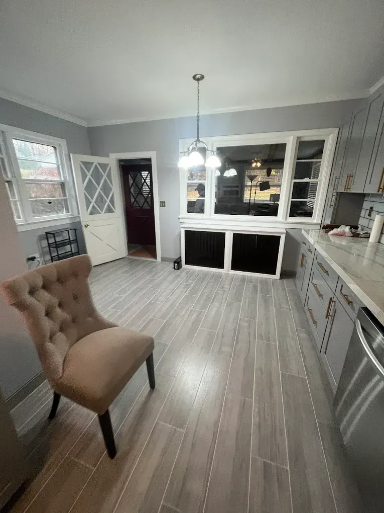 Empty gray kitchen with white cabinets, pendant light, and a tan upholstered chair near the window.