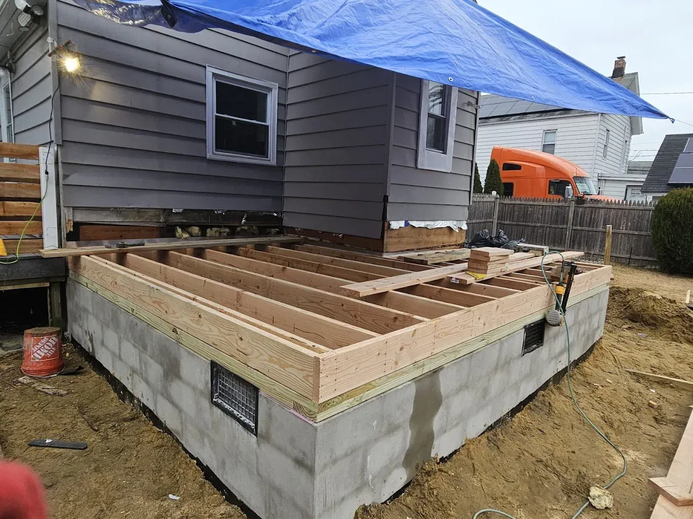House under construction with cinder block foundation and wooden deck framing, covered by a blue tarp