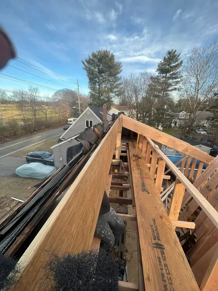 Roof framing under construction, viewed along wooden rafters with a street and houses below.
