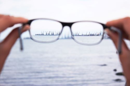 A person is holding a pair of glasses in front of a body of water.