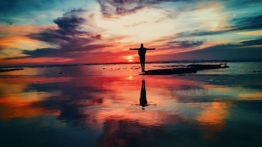 Person with arms outstretched silhouetted against a vibrant sunset reflecting in the water.
