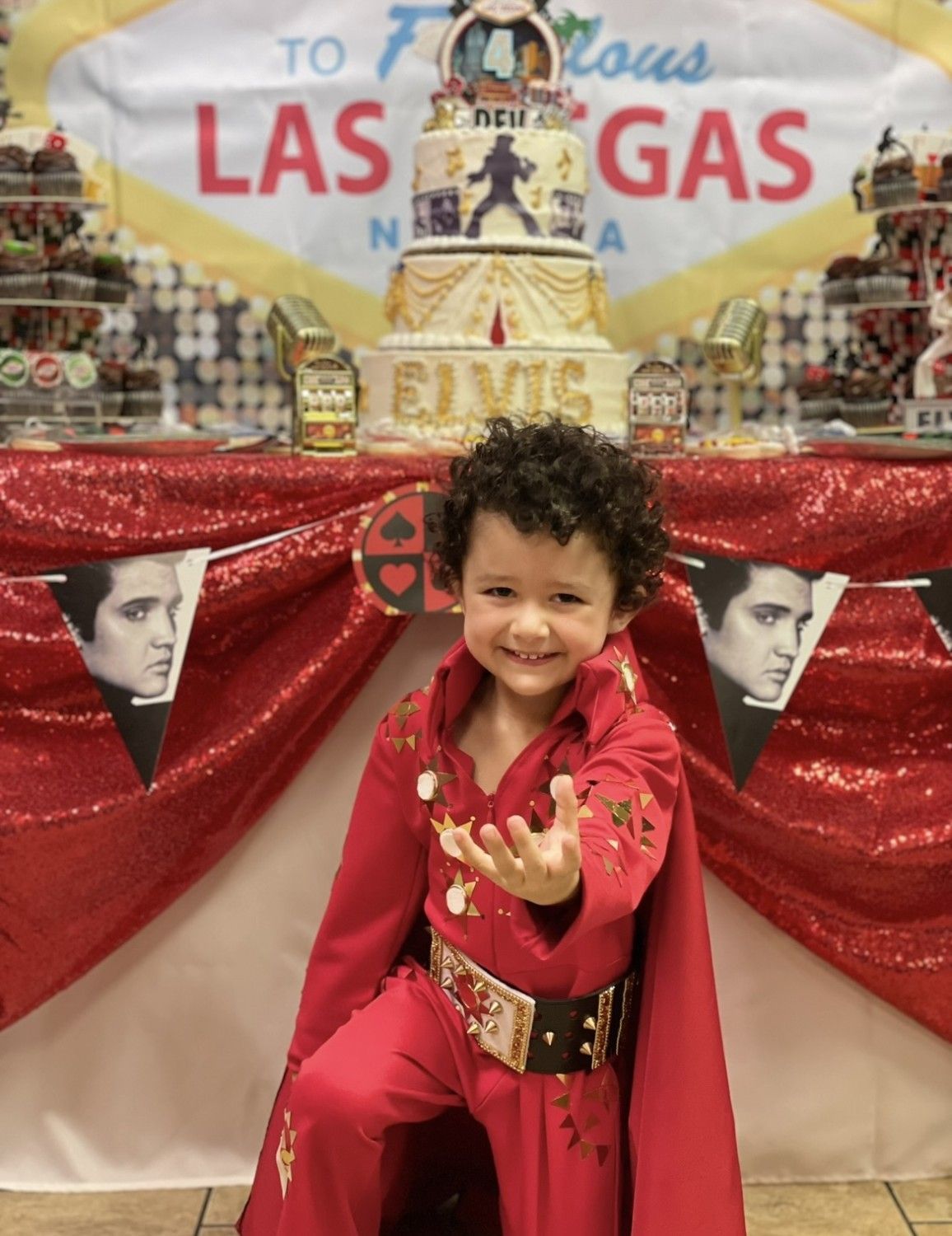 Young child in Elvis costume, smiling, in front of a Las Vegas-themed cake and decorations.