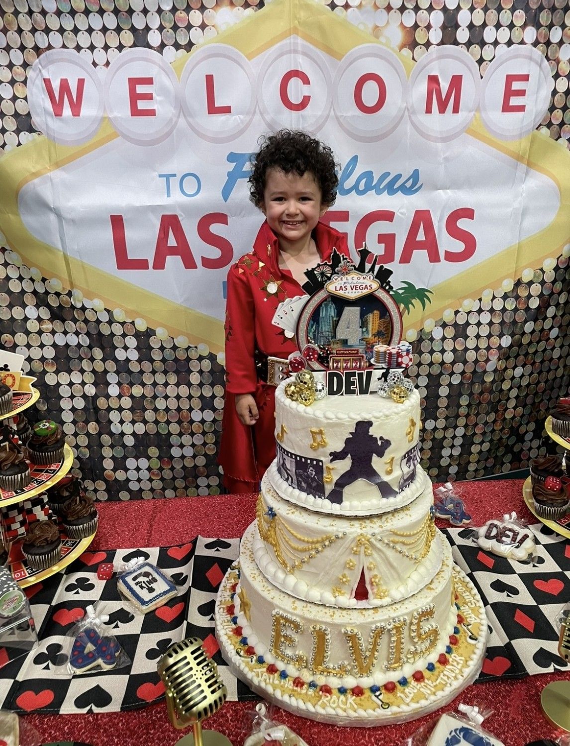 Boy dressed as Elvis poses by a three-tiered cake with an Elvis silhouette, in front of a Vegas-themed backdrop.