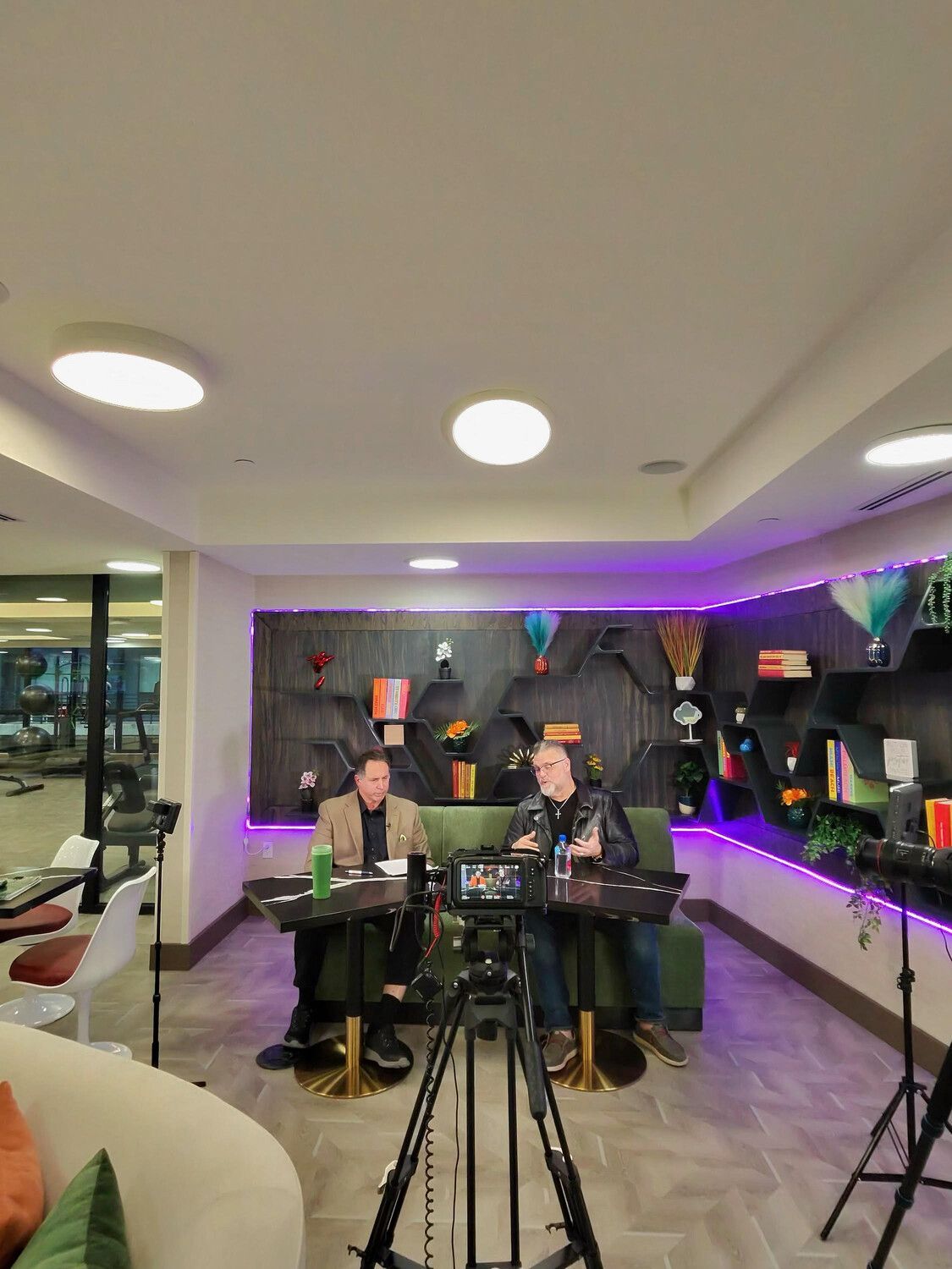 Two men seated at tables, recording a podcast in a studio with a gym visible in the background.