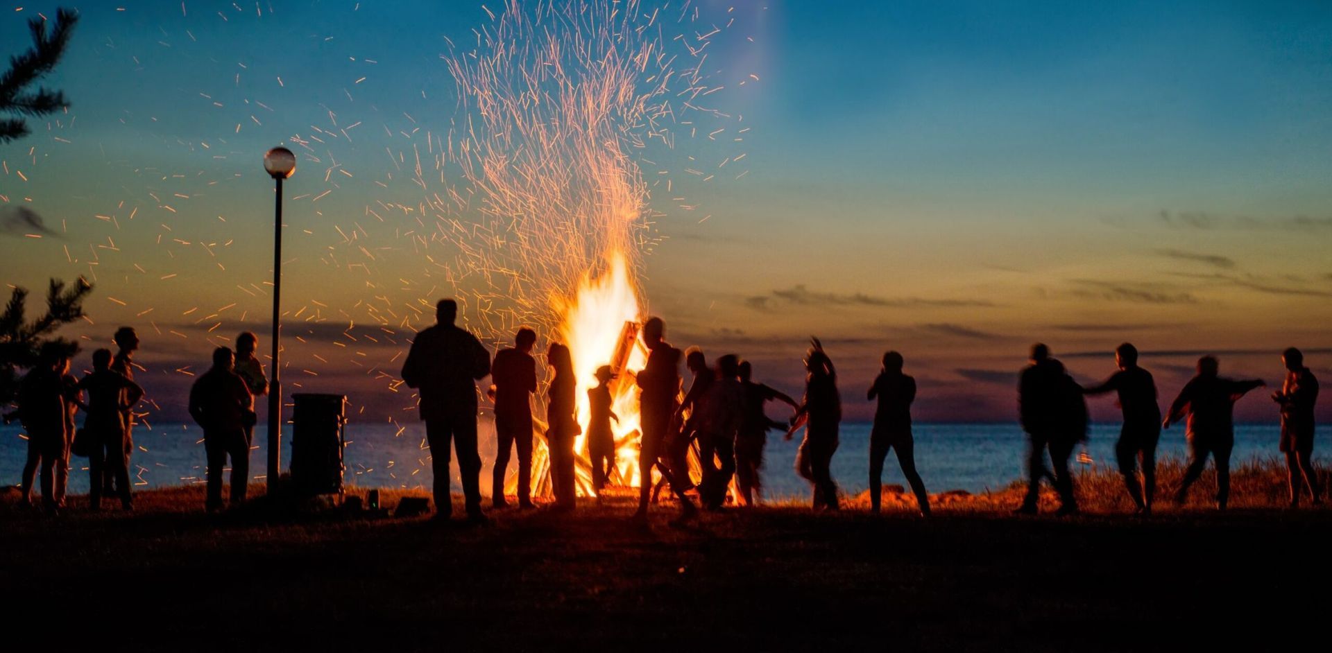 Silhouetted people gather around a large bonfire on a beach at dusk. Sparks fly.