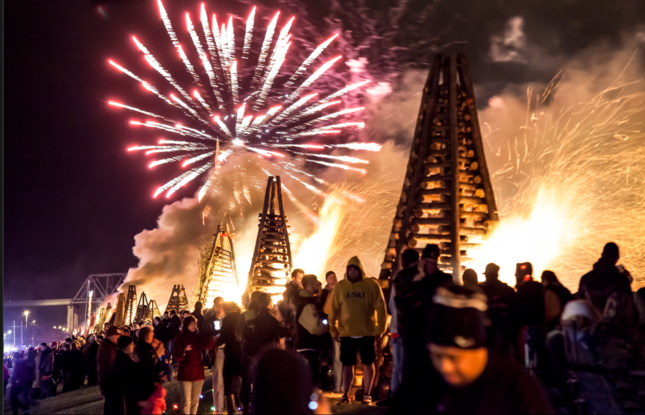 Fireworks erupt above a crowd gathered near burning structures at night.