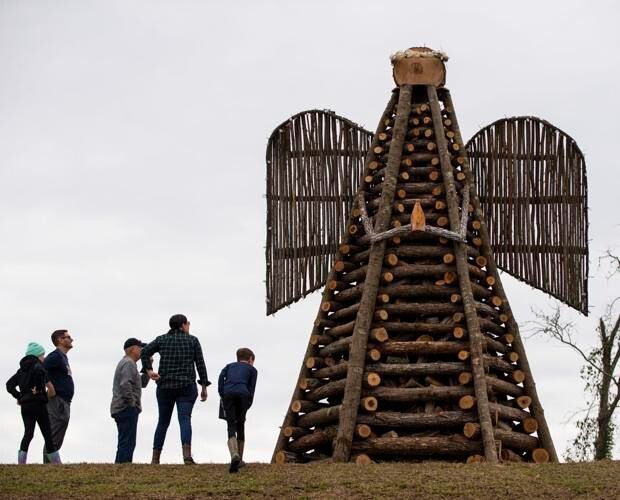 People looking at a large wooden angel sculpture with outstretched wings in an outdoor setting.