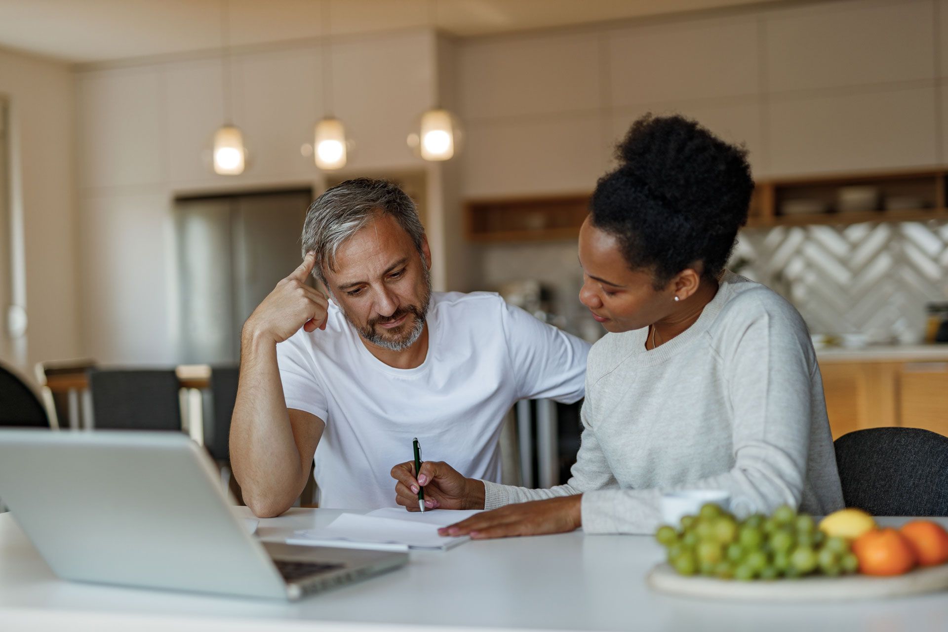 Couple reviewing documents at a table with a laptop, a thoughtful man, and a woman offering assistance, in a modern kitchen.