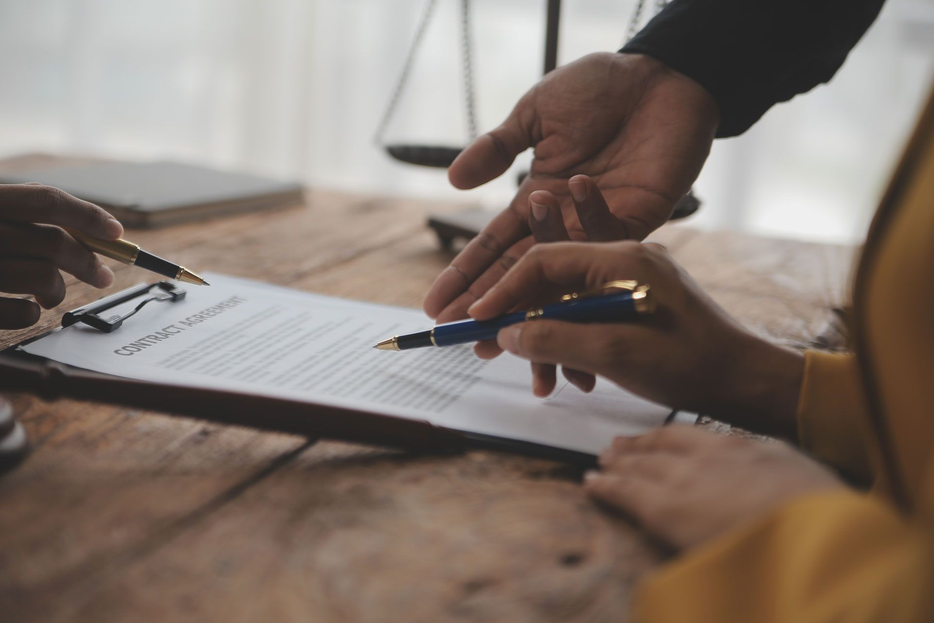 Hands signing document with pen on wooden table, scales of justice in background.