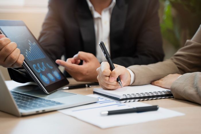 Business meeting with tablet, laptop, and notes on a desk while someone writes with a pen