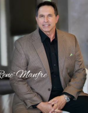 Man in a tan blazer over a black shirt, smiling slightly, sitting indoors.