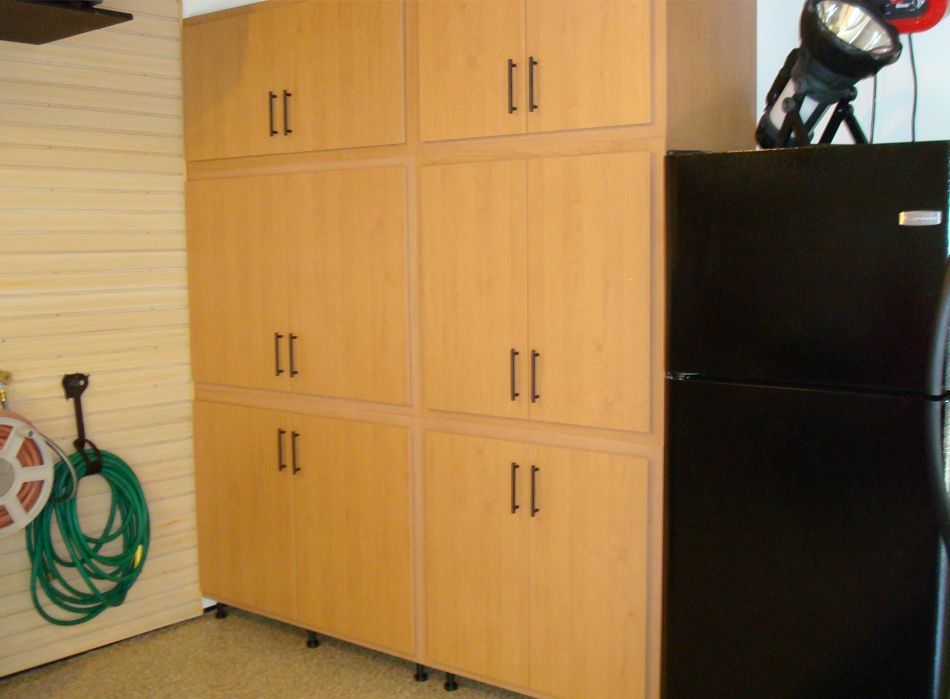 Light-colored storage cabinets and a black refrigerator in a garage. A coiled green hose hangs on the wall.