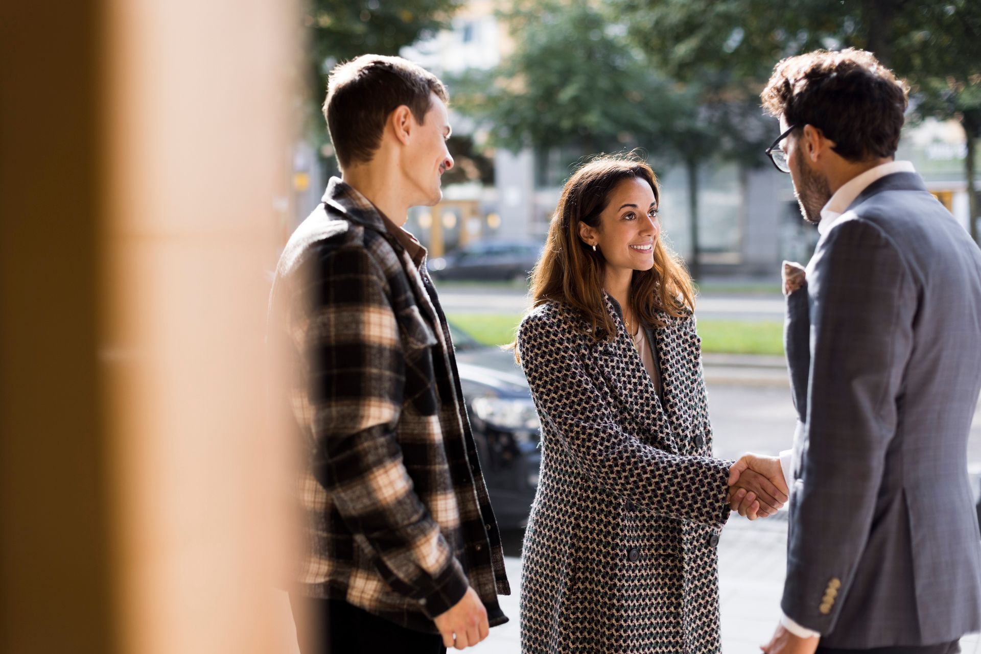 Three people greeting each other outside. A woman shakes hands with a man in a suit, while another man looks on. Sunlight.