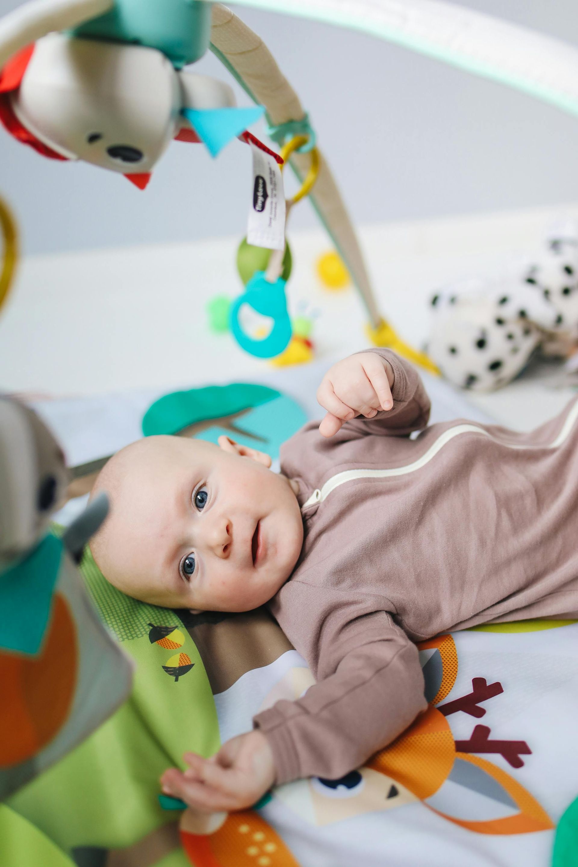 Baby lying on a colorful play mat, reaching toward hanging toys while looking at the camera.
