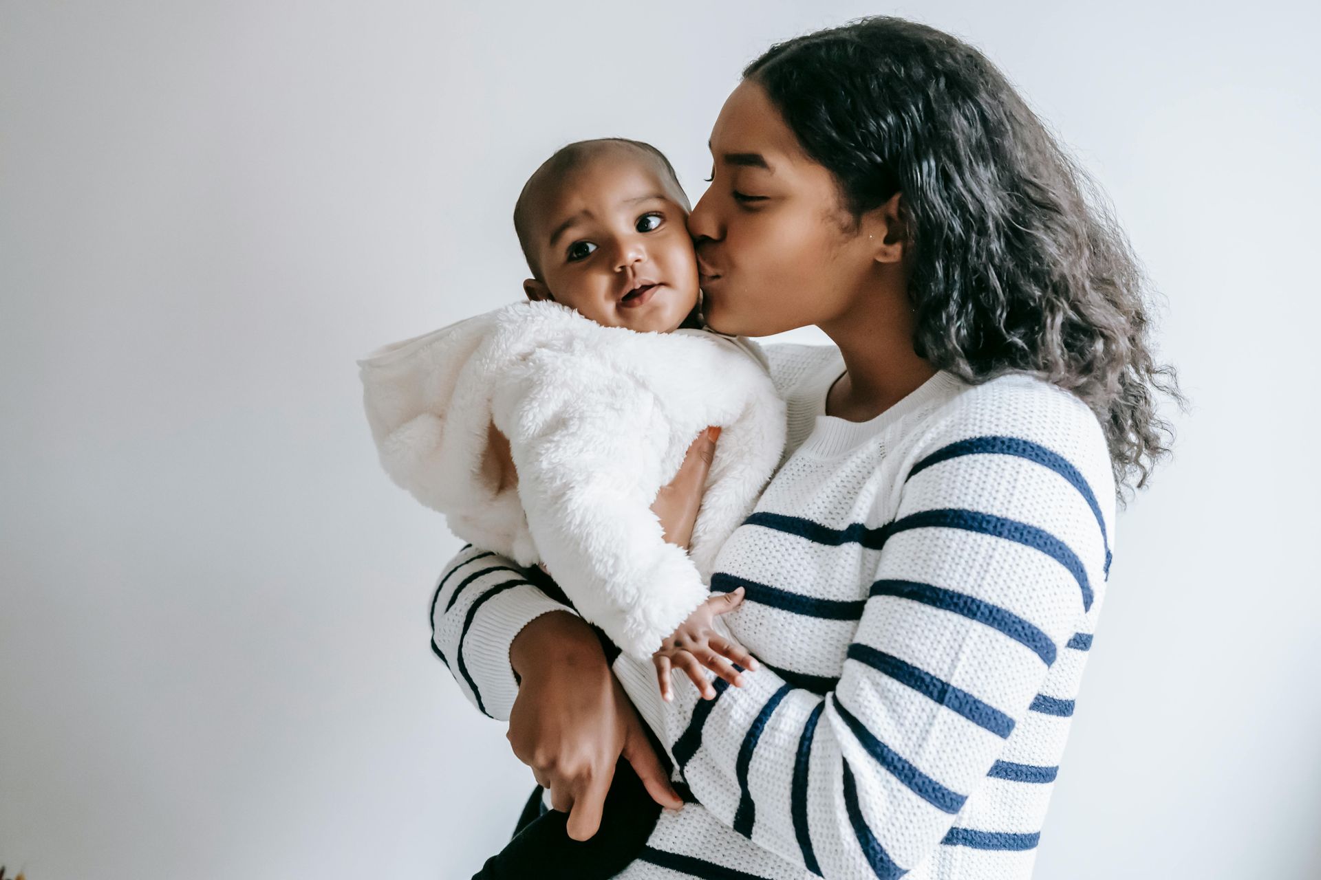 Woman in a striped sweater kissing a baby she is holding against a light background