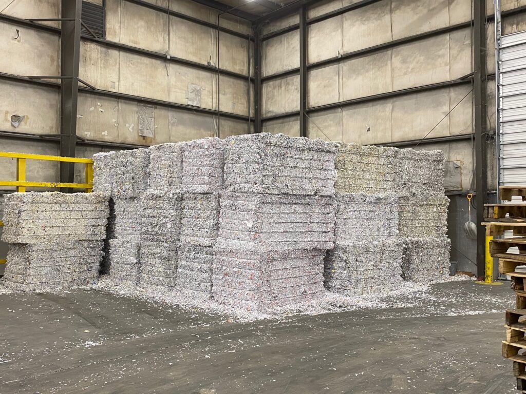 Stacks of shredded paper bales inside a warehouse, on a concrete floor near a loading dock.